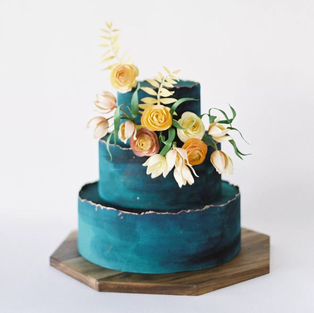A three-tiered wedding cake of ocean blue brushstrokes with metallic trim and sunshine-hued blooms