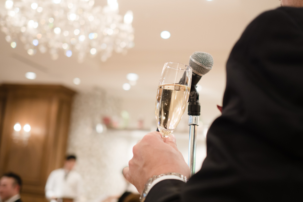 Man in tuxedo at microphone giving speech with champagne glass in hand