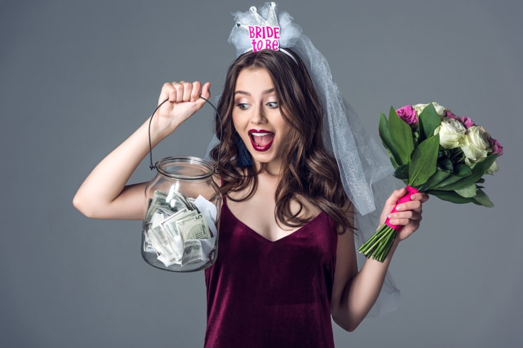 shocked future bride in veil for bachelorette party with flowers looking at jar of money