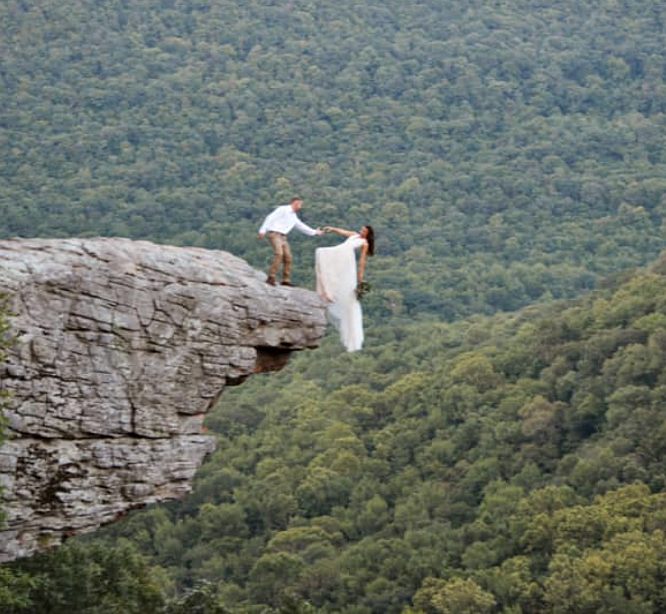 Bride dangles off cliff while groom reaches his hand out to her for an epic wedding photo