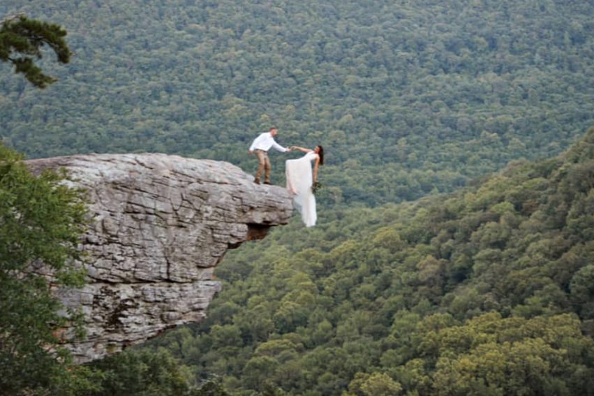 Bride dangles off cliff while groom reaches his hand out to her for an epic wedding photo