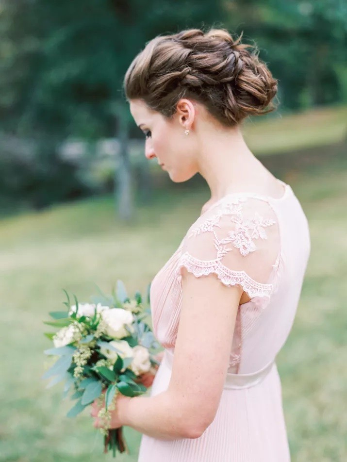 Bridesmaid wearing romantic pale pink dress with lace cap sleeves looks at bouquet for photo.
